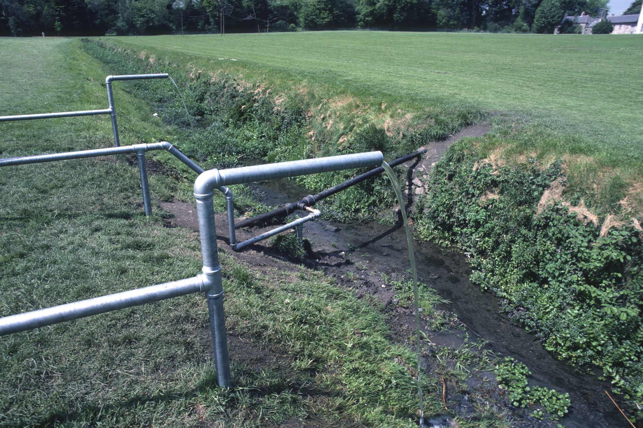 Water flowing from two galvanized pipes and dropping down into a muddy ditch
