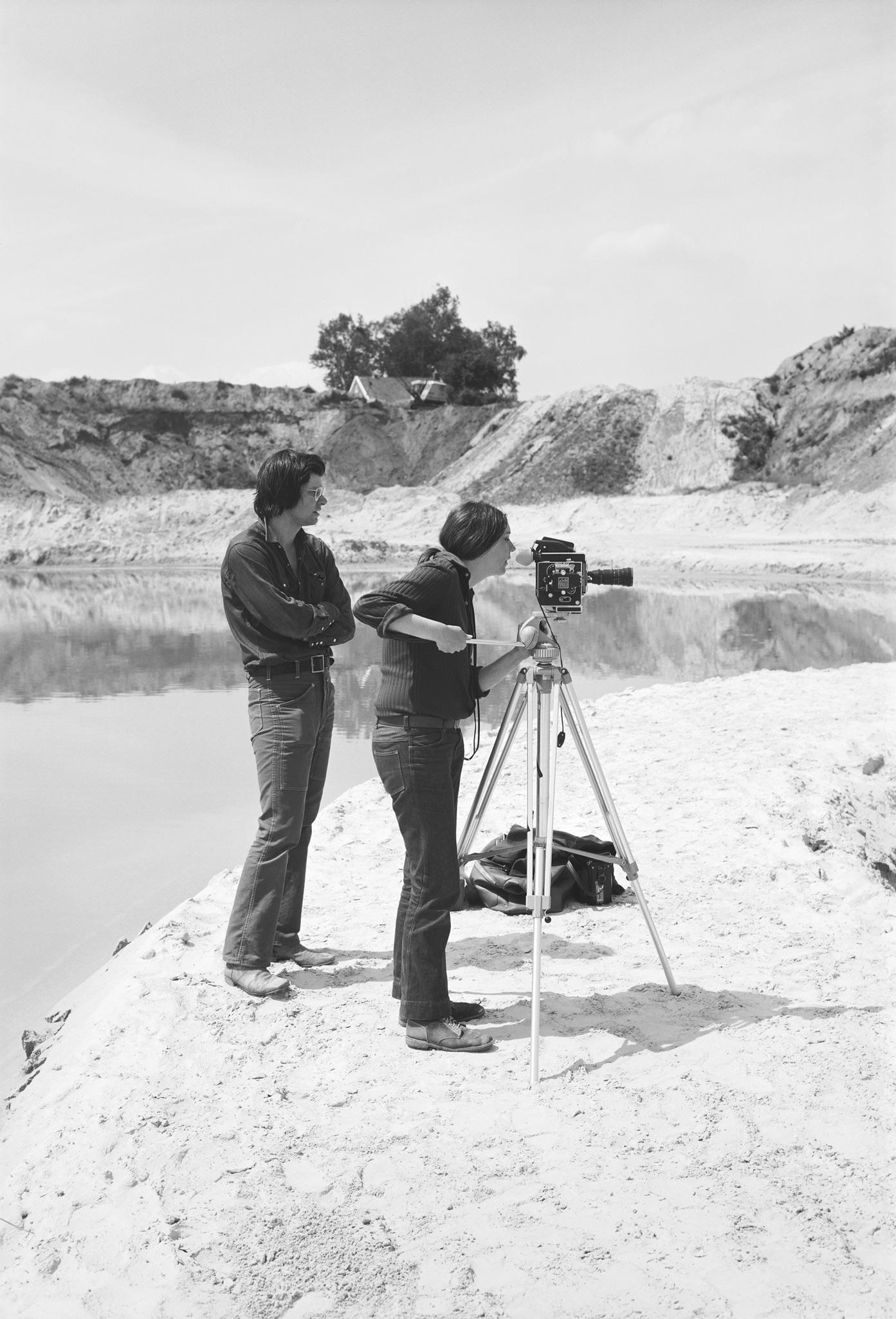 two people stand on a sandy bank looking to the right and one is looking through a film camera on a tripod