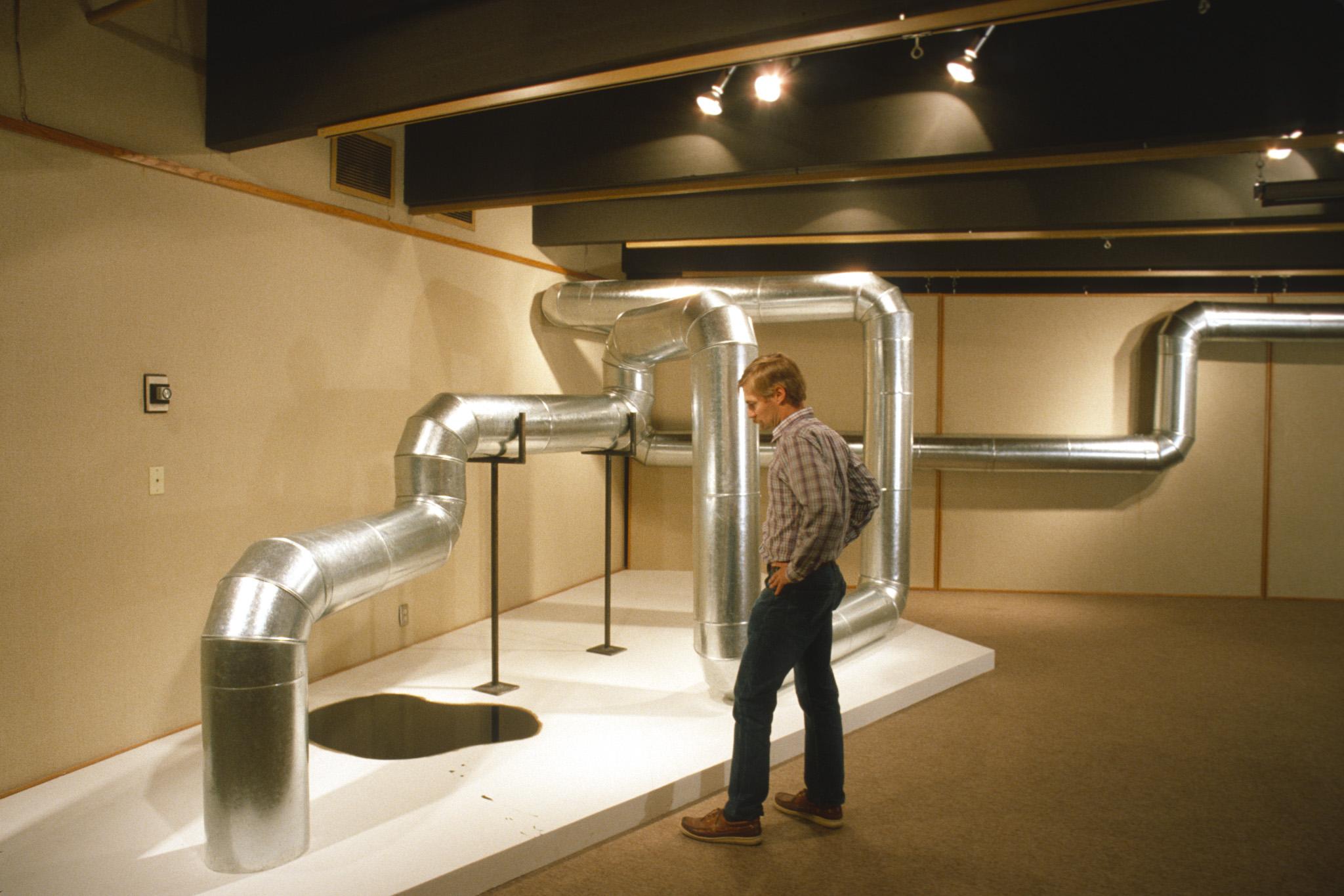 a person stands next to a sculpture made of steel duct that zig zags through the air and along the walls in a basement gallery space