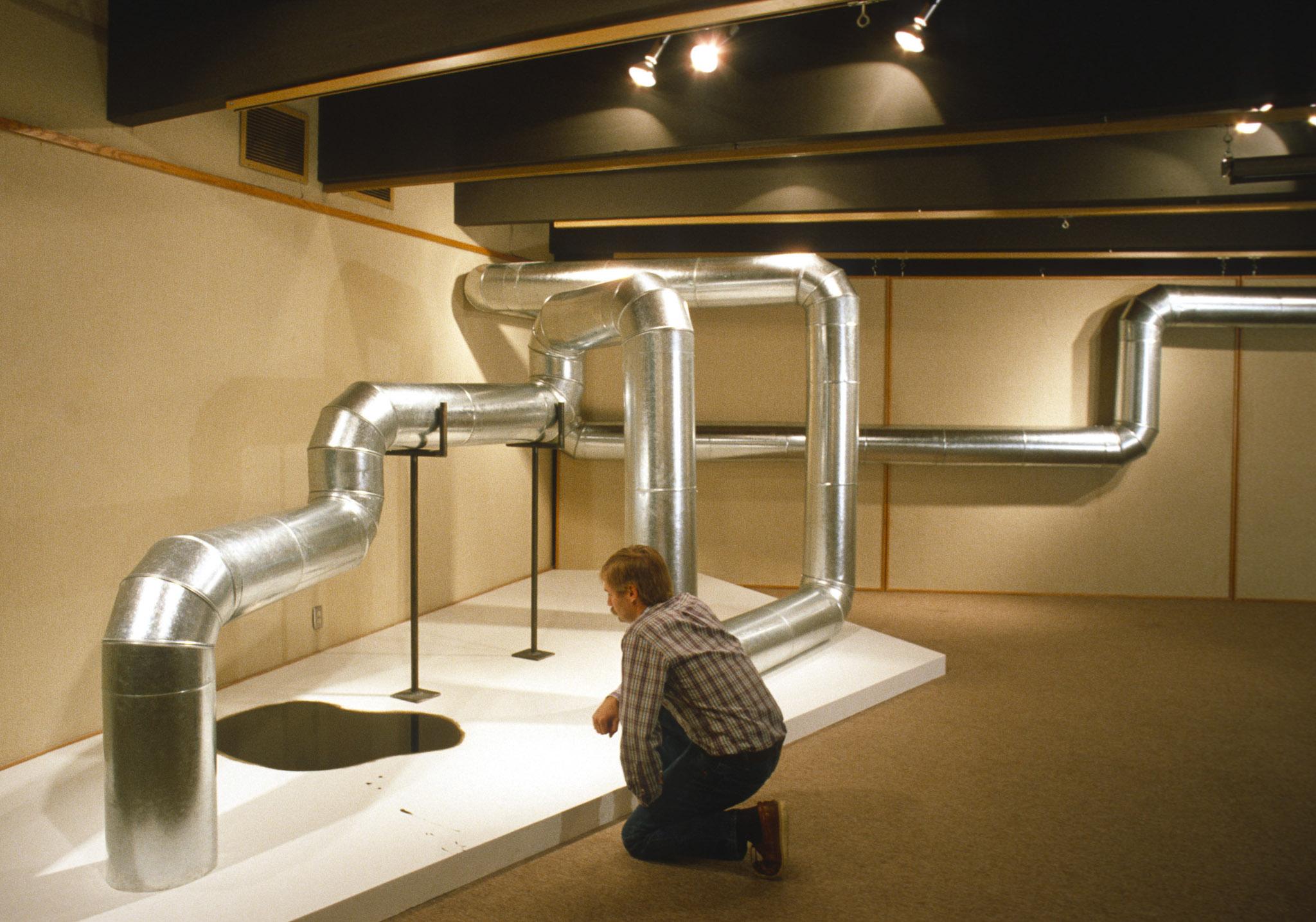 a person kneels next to a sculpture made of steel duct that zig zags through the air and along the walls in a basement gallery space