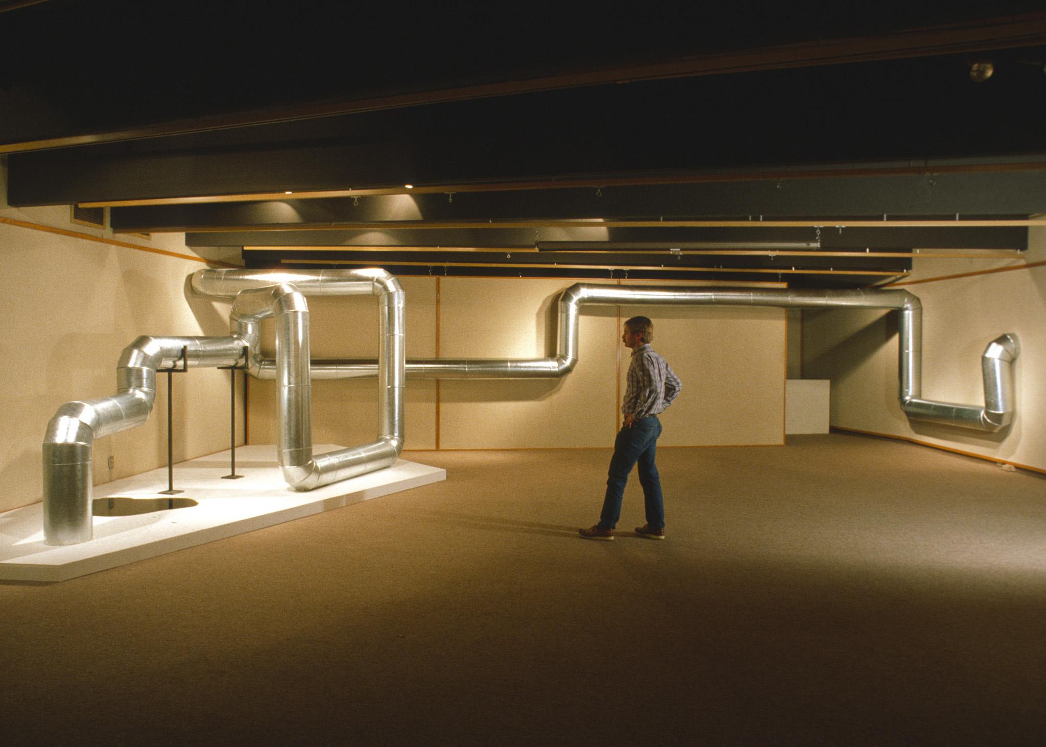 a person stands next to a sculpture made of steel duct that zig zags through the air and along the walls in a basement gallery space