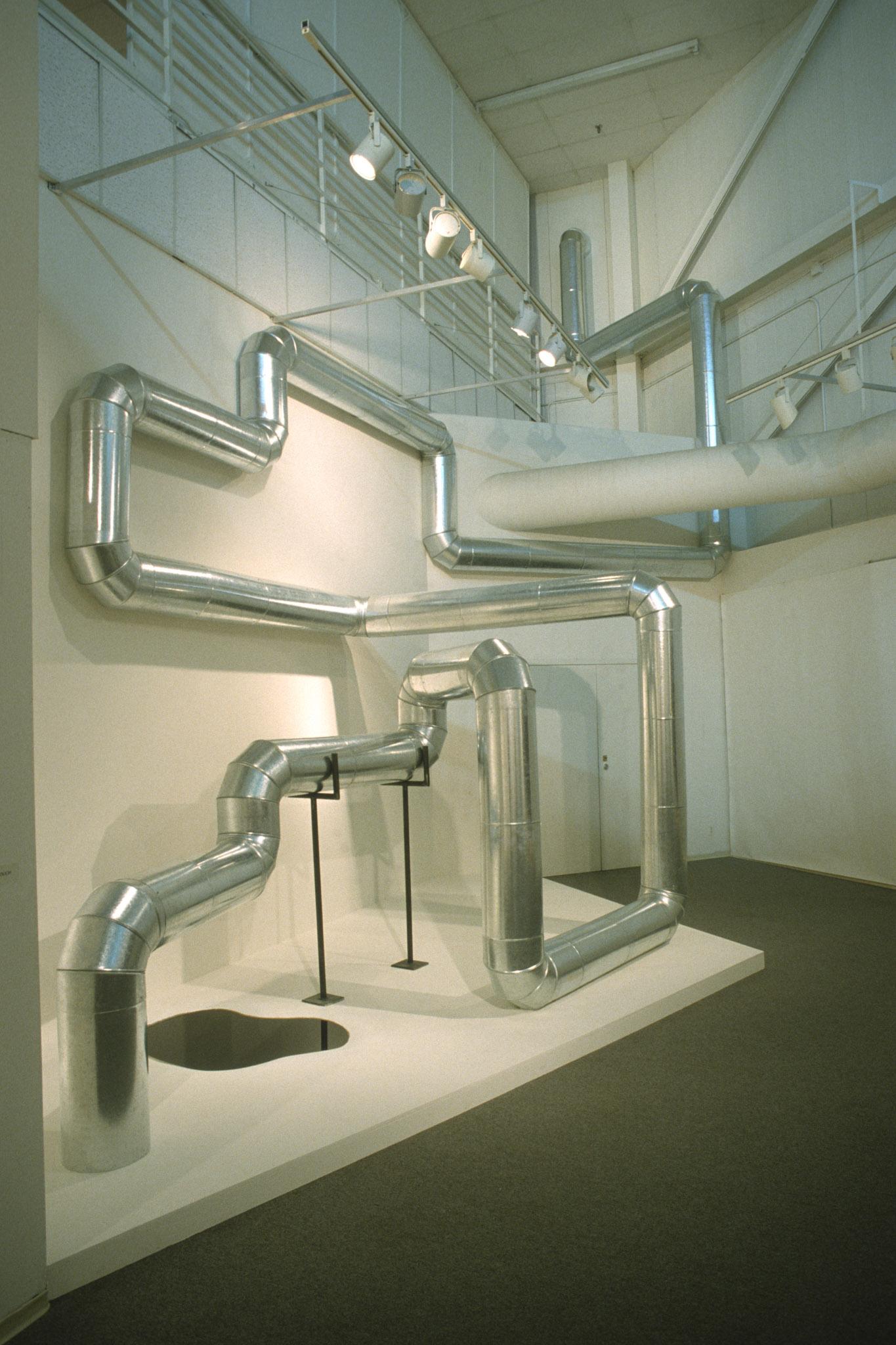 steel duct zig zags through the air and along the walls inside a white gallery space. A small puddle of oil has pooled on the floor.