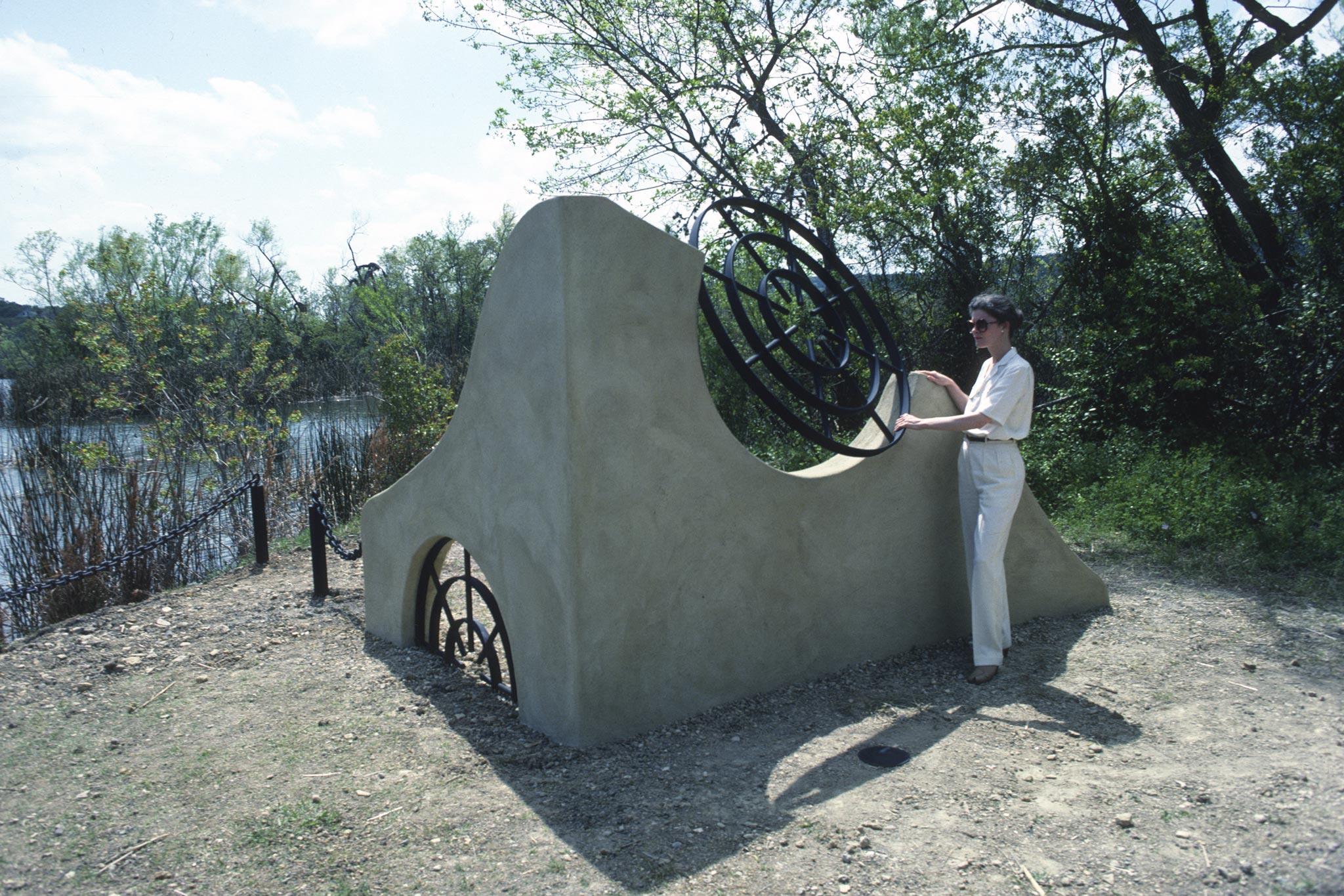 person stands next to a short undulating stucco wall with two round metal grates incorporated into the structure built on a lush lake shoreline
