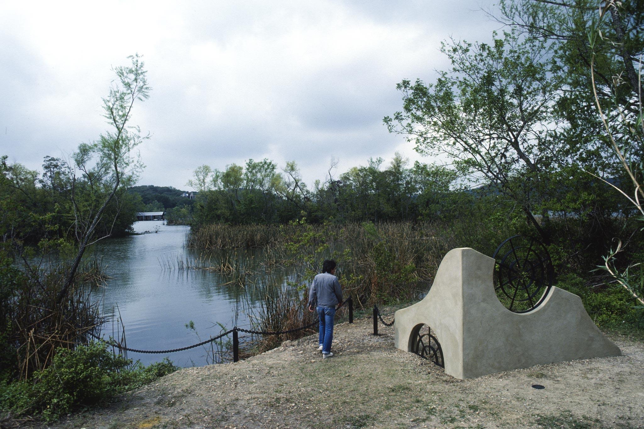 person stands next to a short undulating stucco wall with two round metal grates incorporated into the structure built on a lush lake shoreline