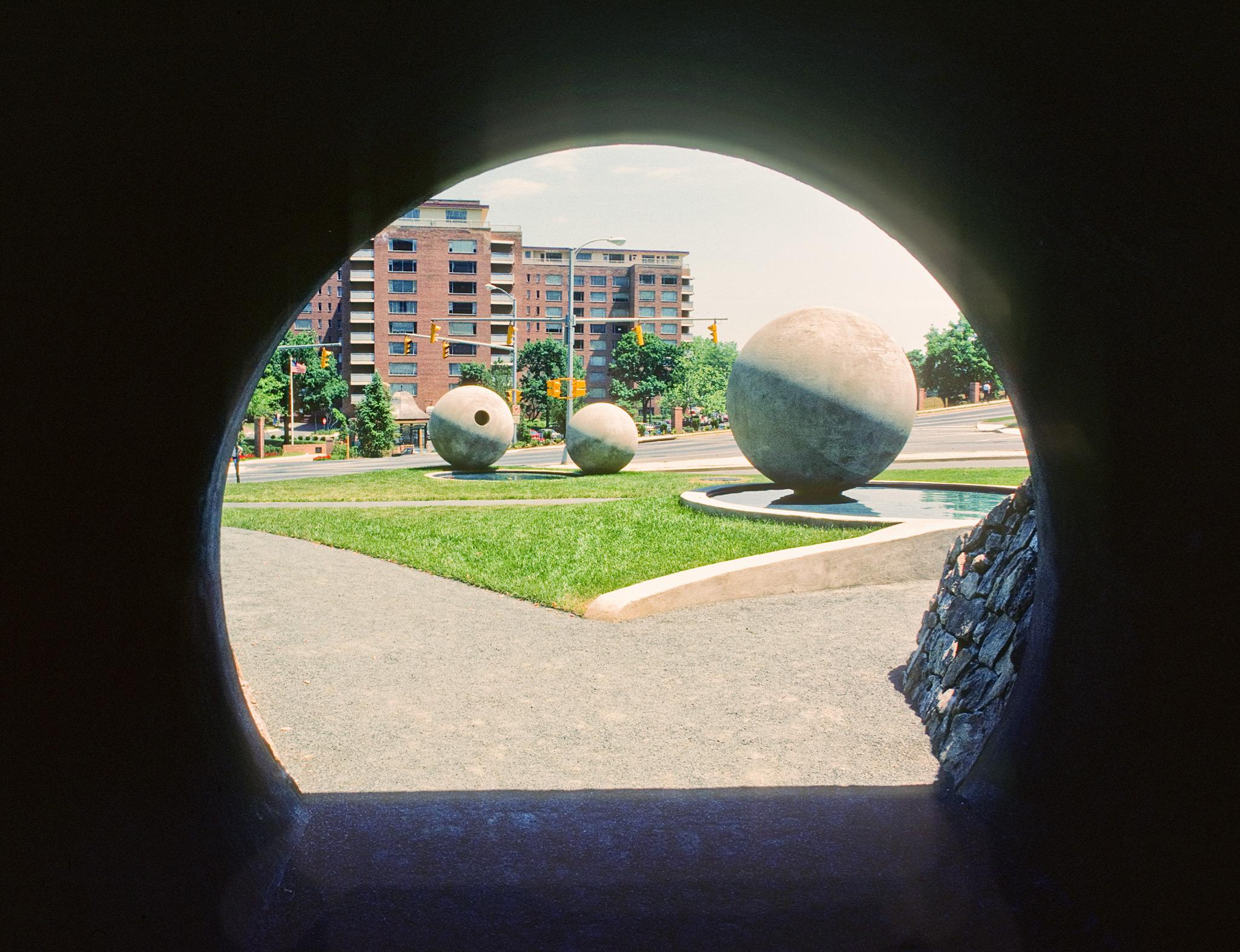 looking out of a circular tunnel to three large concrete spheres in a city park
