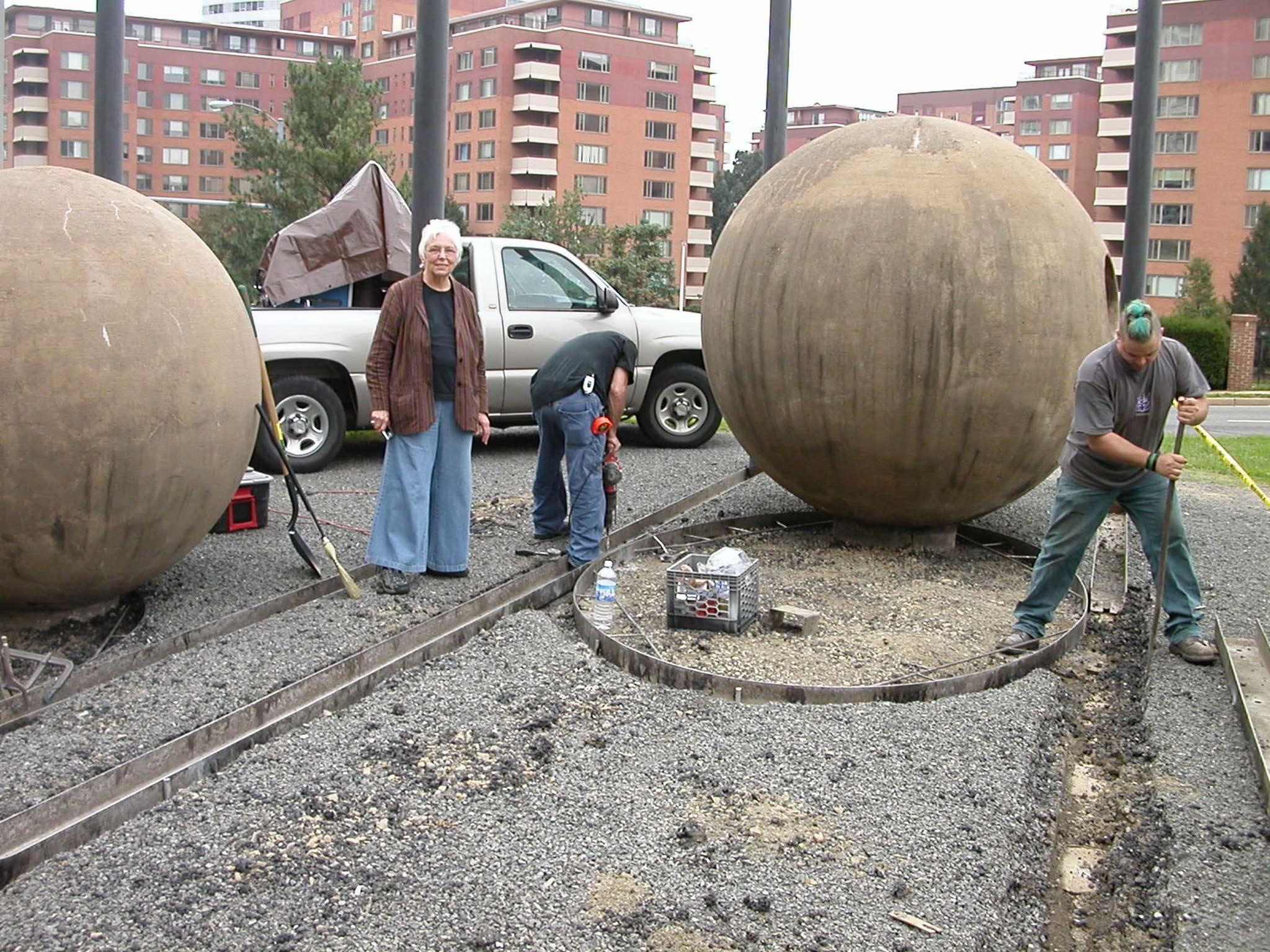 two figures working and one figure looking at the camera at a construction site, renovating large concrete spheres.
