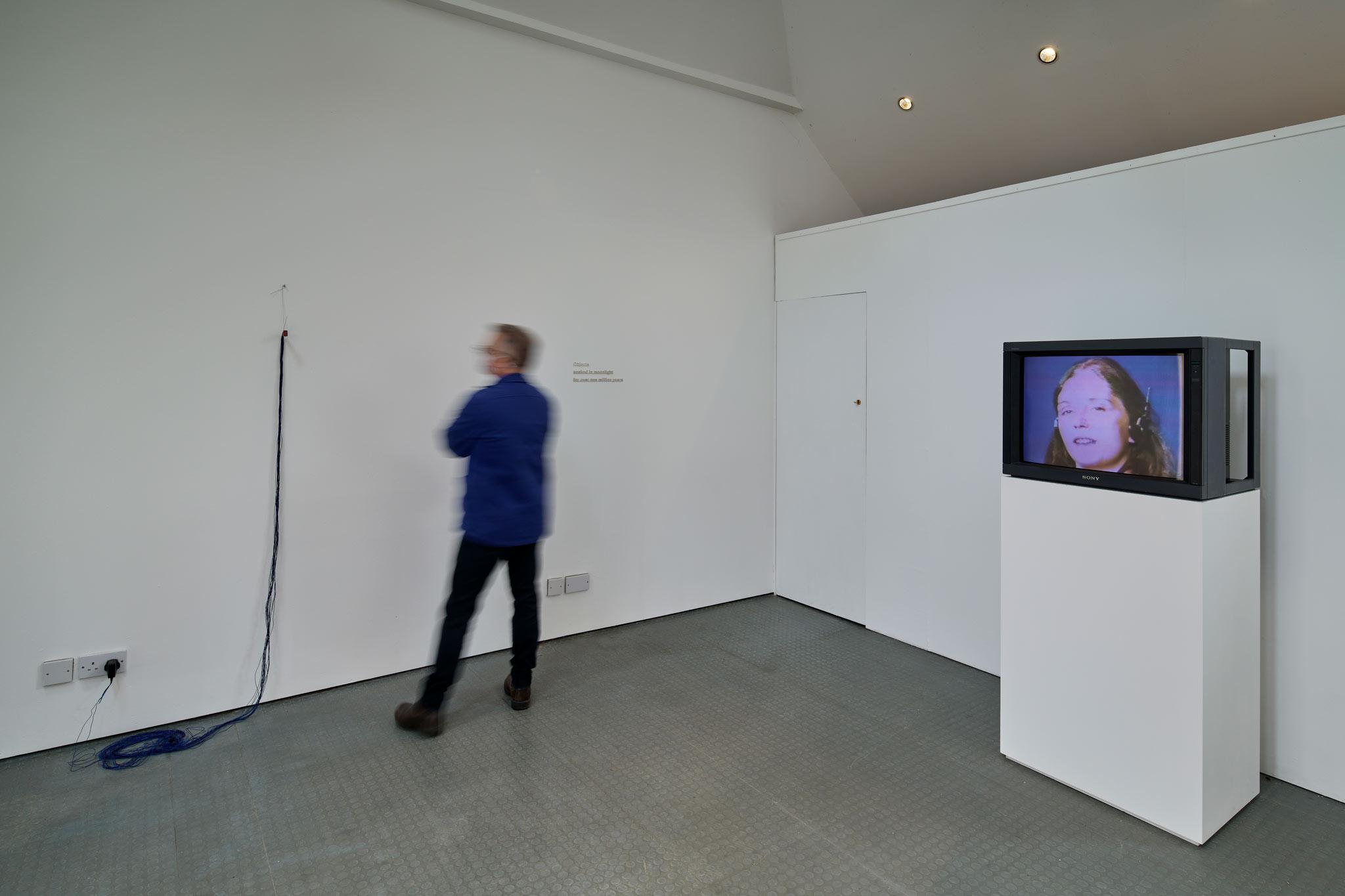 a person standing in a gallery space with a monitor on the right side and a wire sculpture on the left