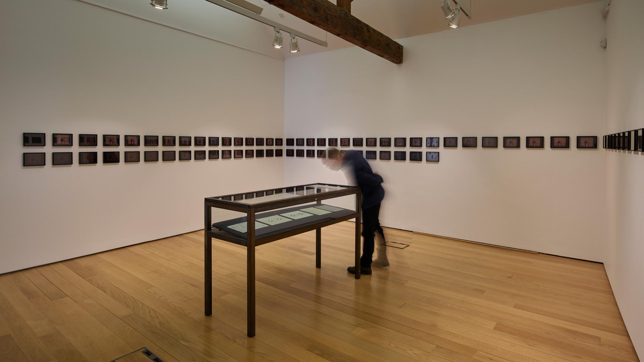 a gallery space with small dark photographs in a line wrapping around the walls and a display case in the center of the room with a person looking down into it.