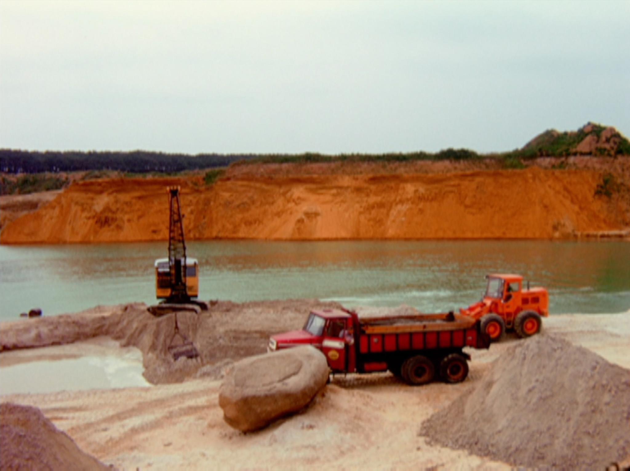 trucks and dozers moving earth in a sand quarry with a lake behind them