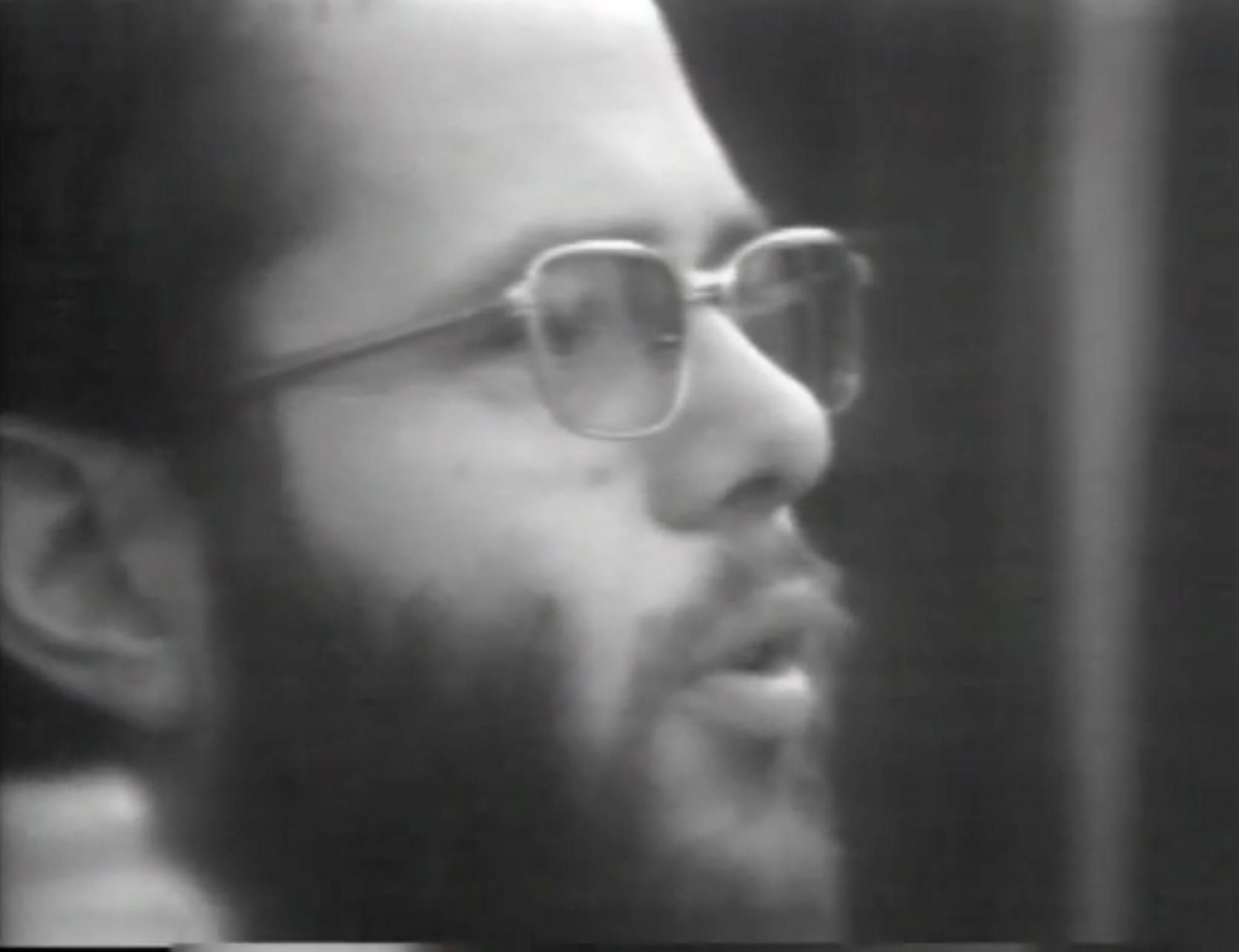 Black and white image of a man with a beard and glasses talking looking to the right.