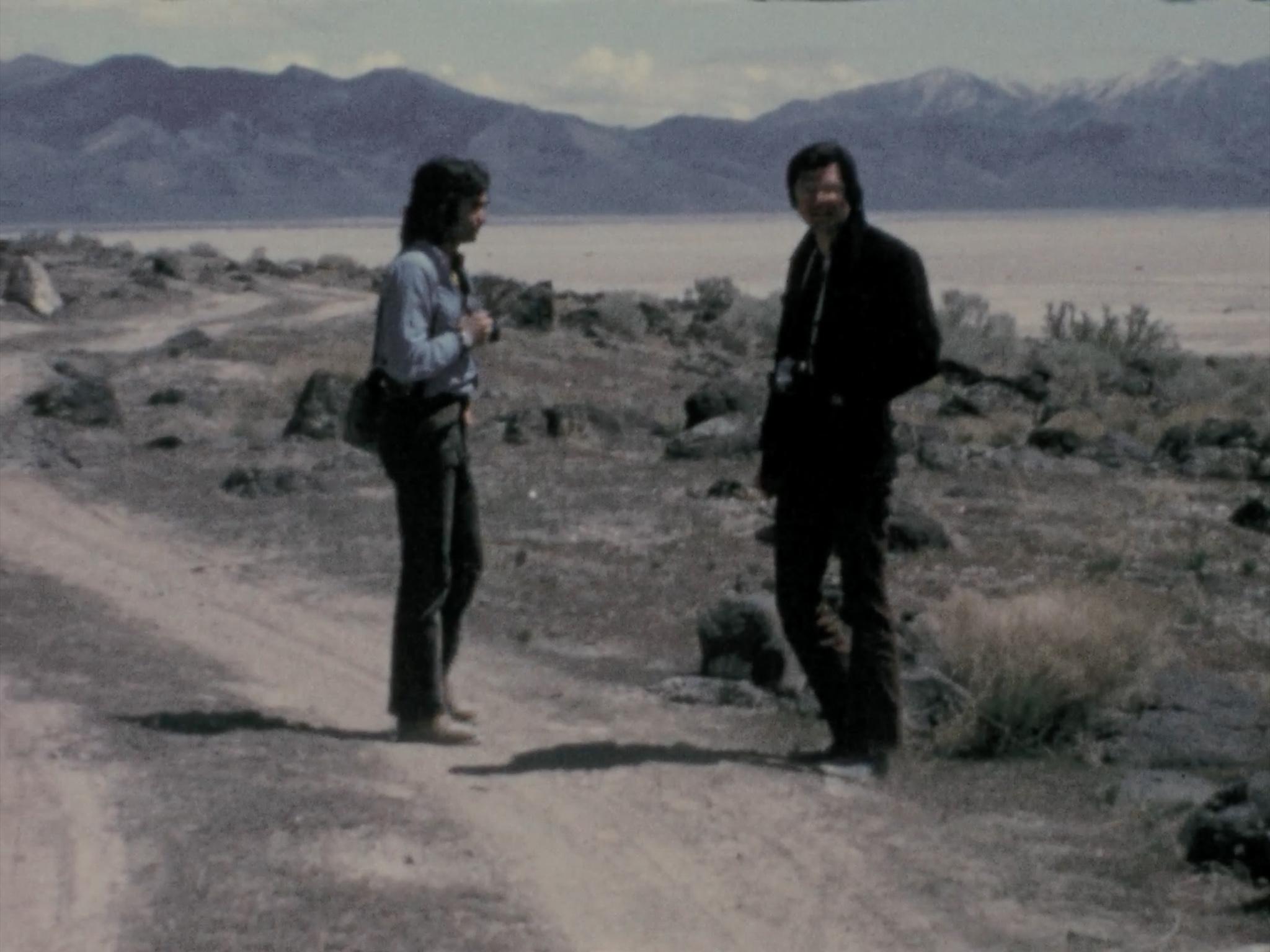 Two figures standing on a dirt road with mountains in the background