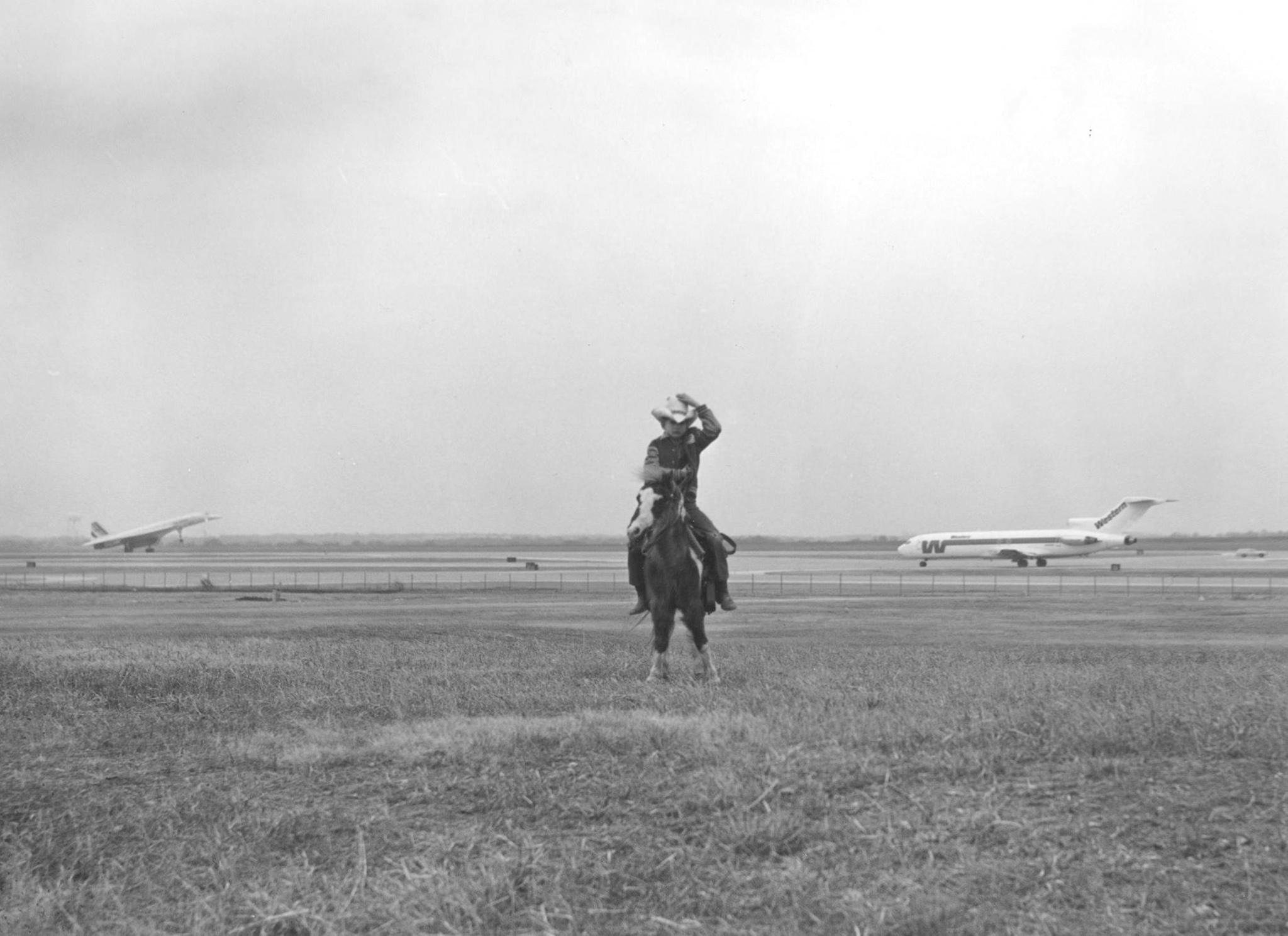 a young boy riding a pony stands in the middle of a field with planes taking off in the background