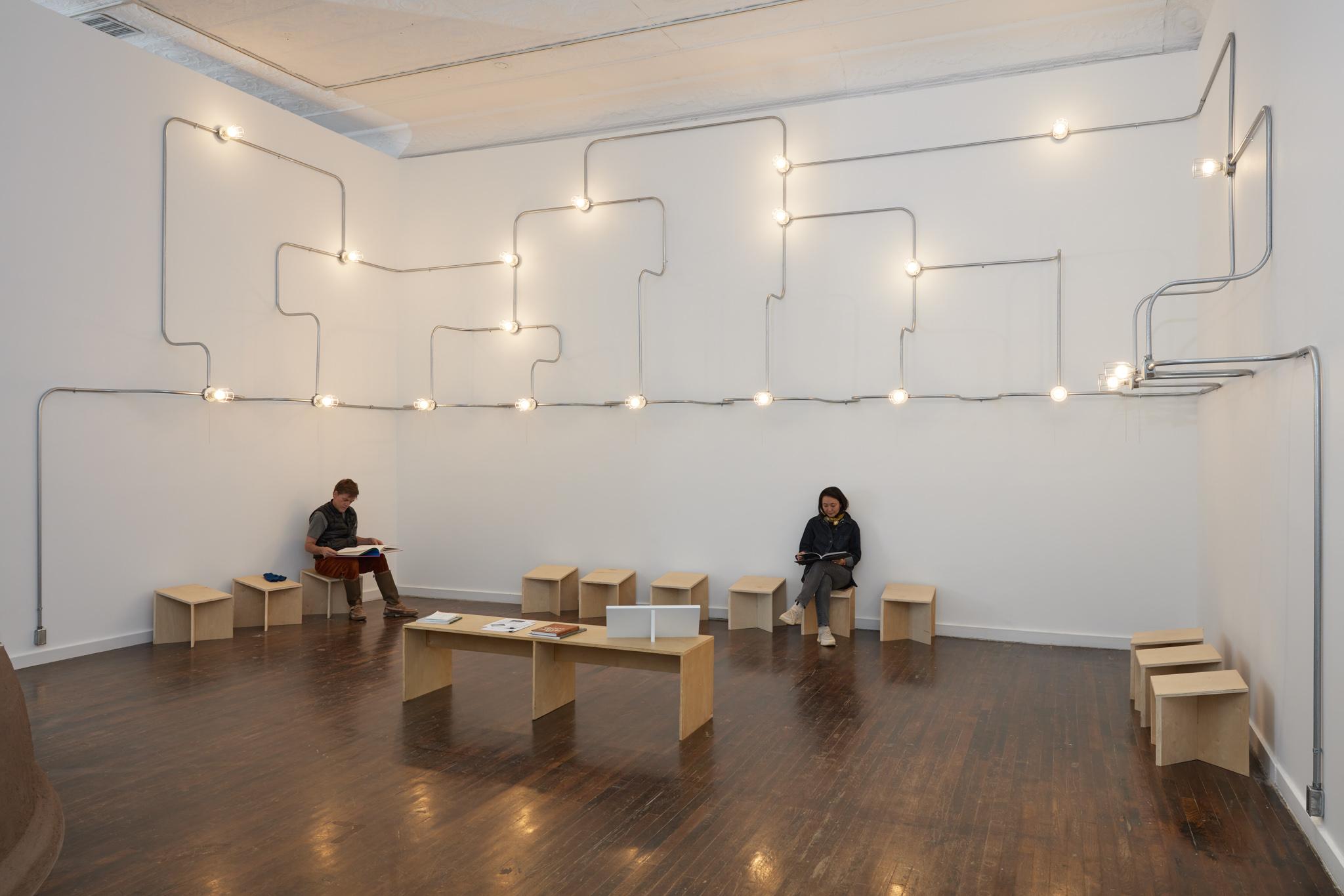 A network of conduit a lightbulbs on three walls above two people sitting on stools