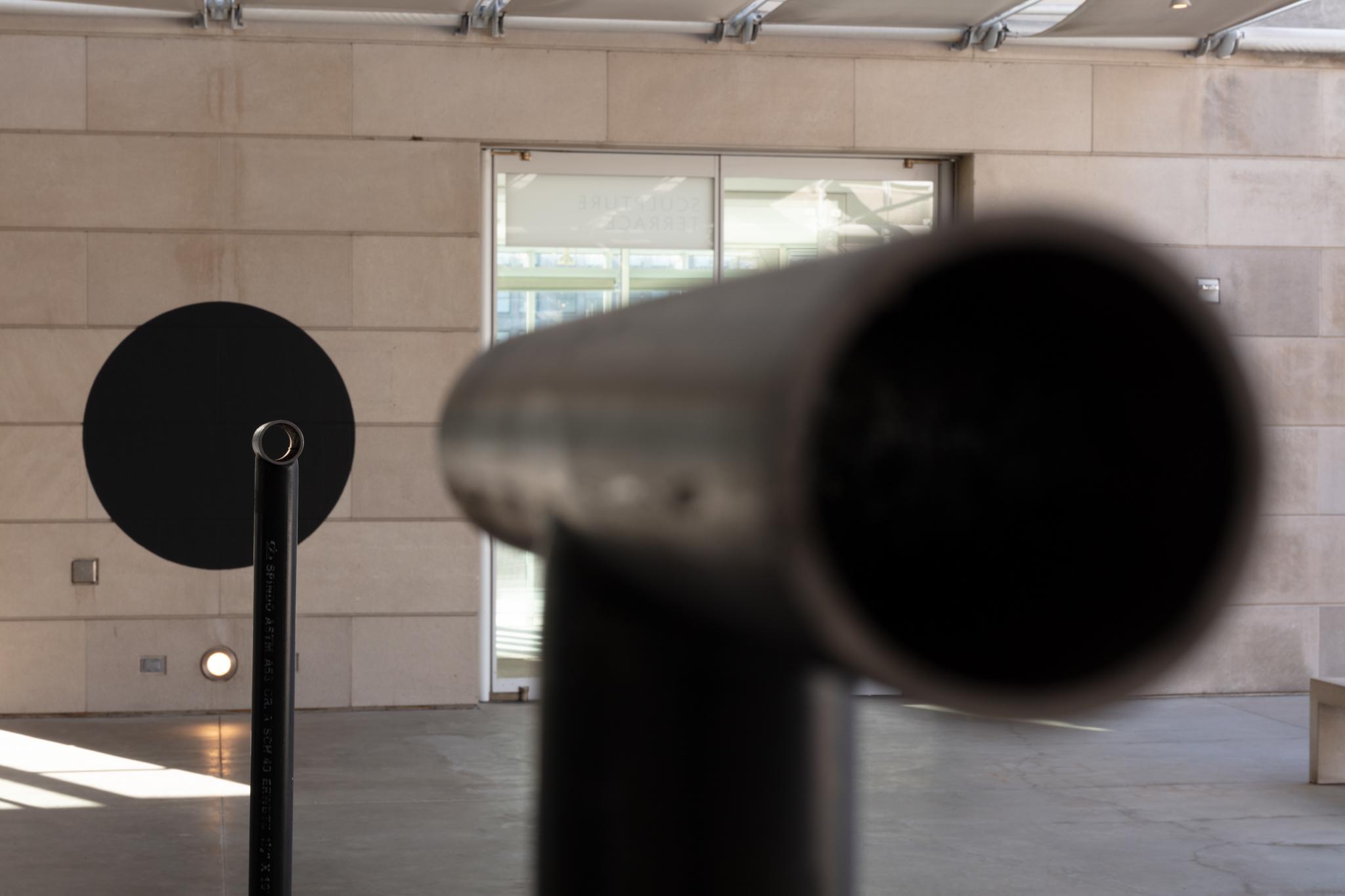 Two Locator sculptures installed on an outdoor terrace pointing toward a large painted black circle on the wall