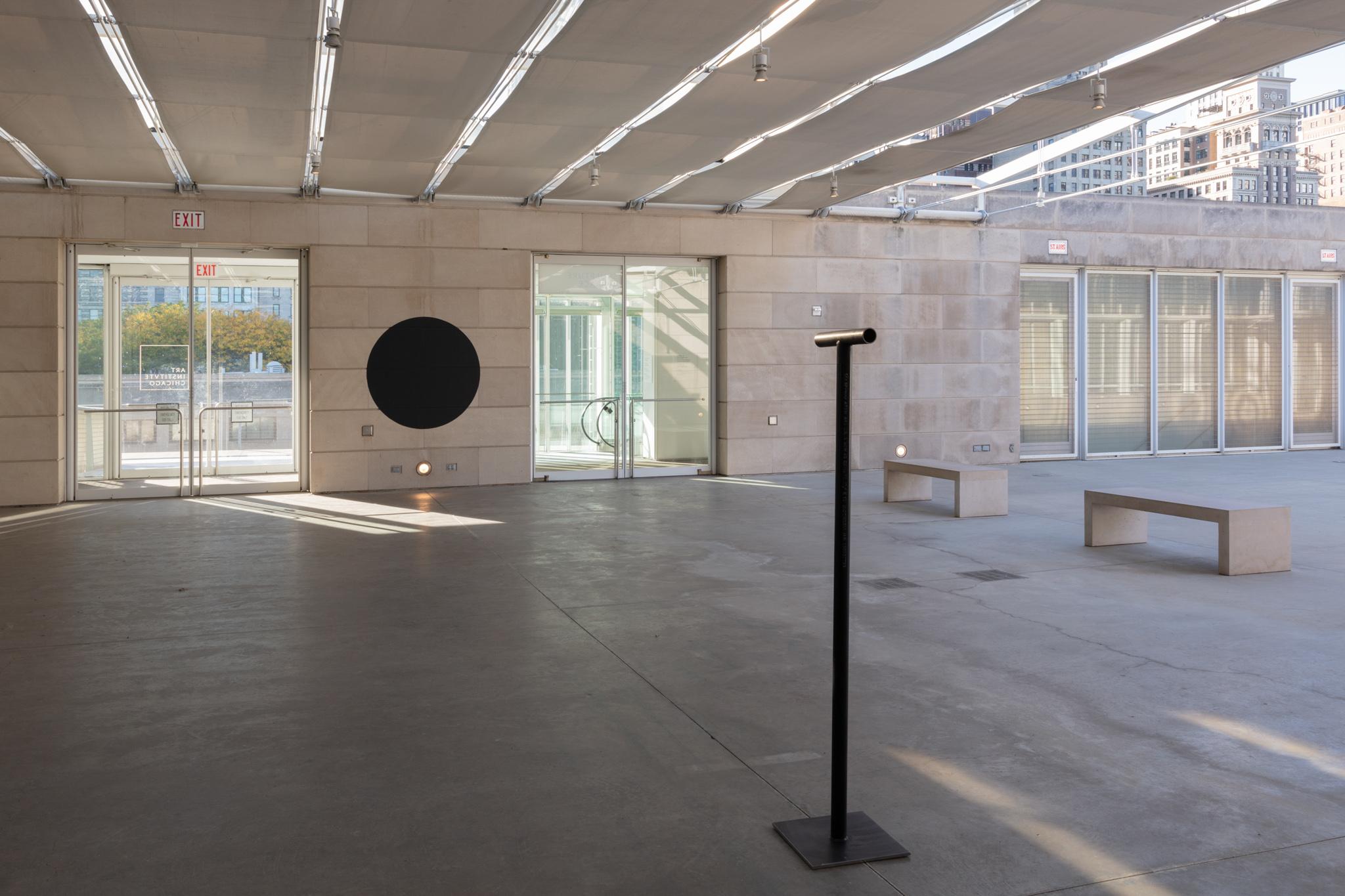 A locator sculpture installed on an outdoor terrace pointing toward a large painted black circle on the wall