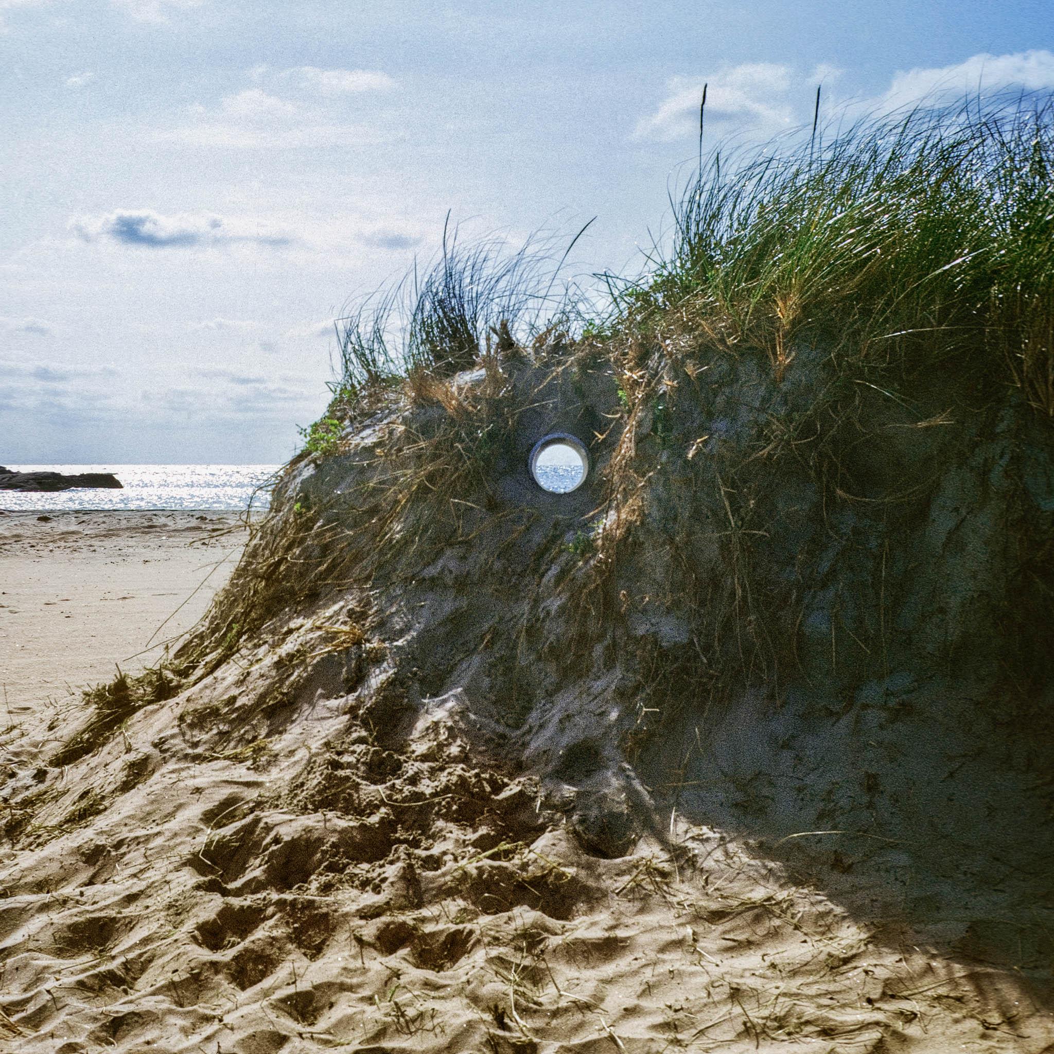 A circular pipe inserted through a sand dune framing a view of ocean and landscape horizons