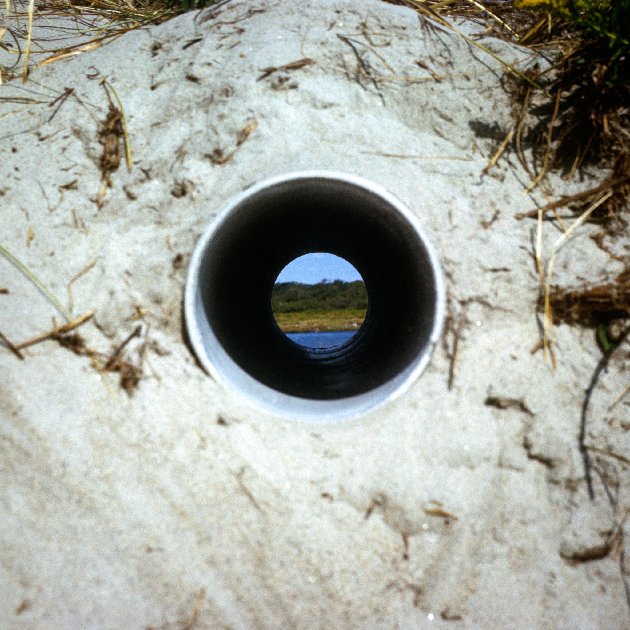 A circular pipe inserted through a sand dune framing a view of ocean and landscape horizons