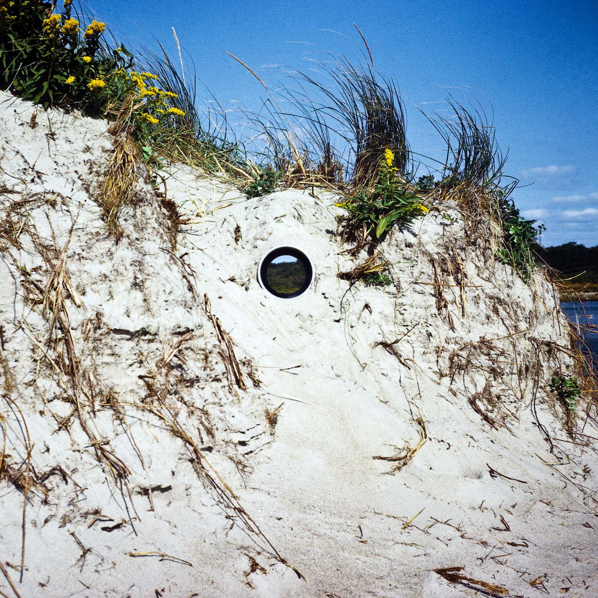 A circular pipe inserted through a sand dune framing a view of ocean and landscape horizons