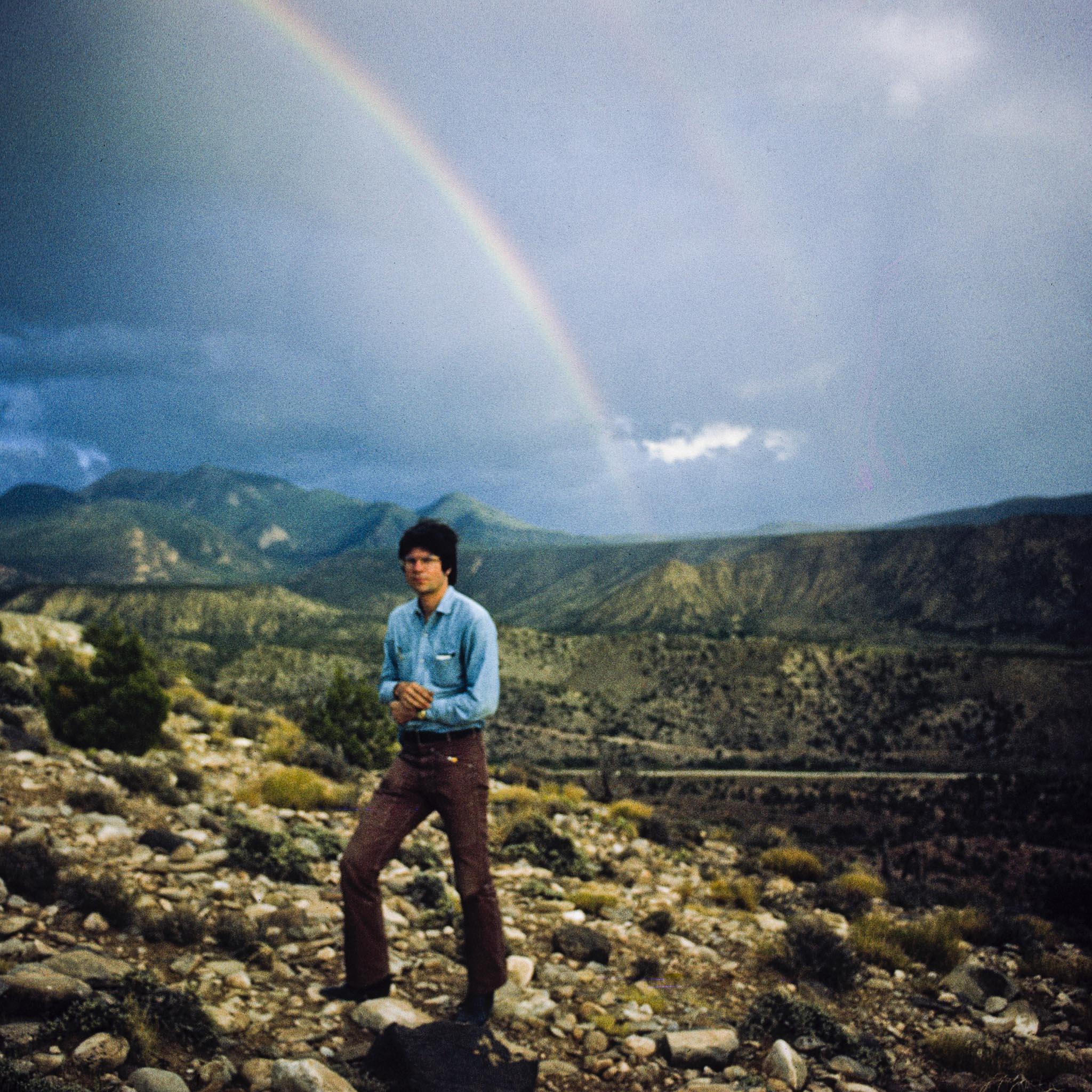 Robert Smithson standing on a hill with a large mountain landscape and rainbow in the background