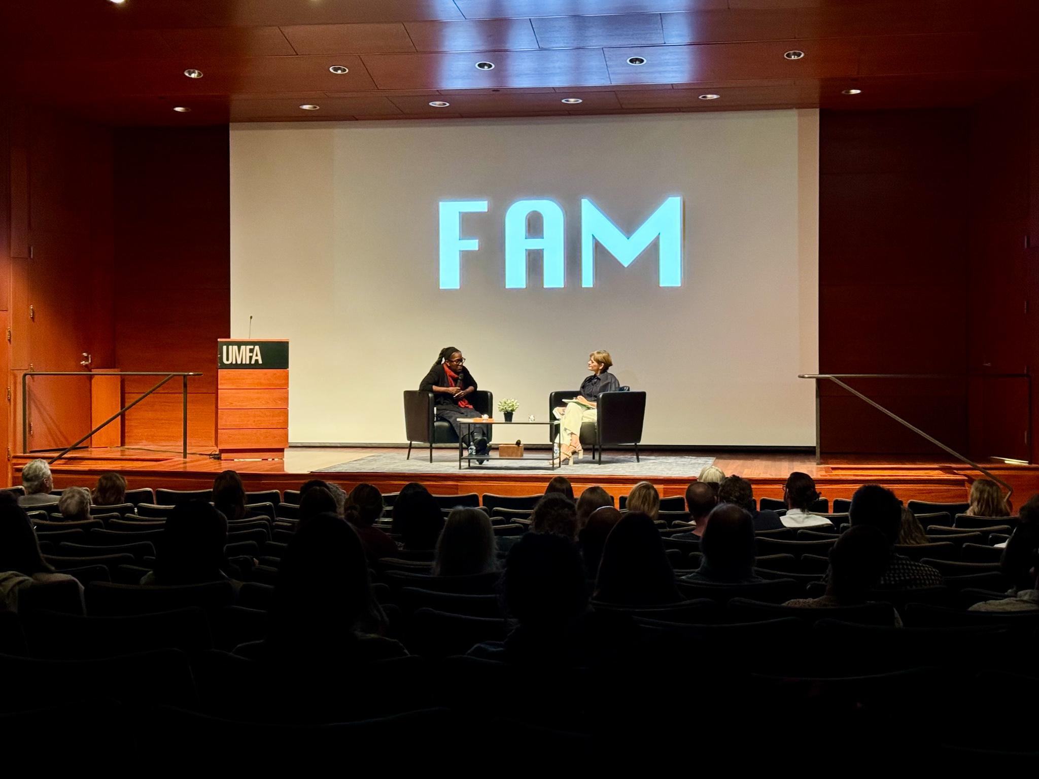 two people sitting on a stage with a projected image behind them that says "FAM" and with a crowd in the foreground