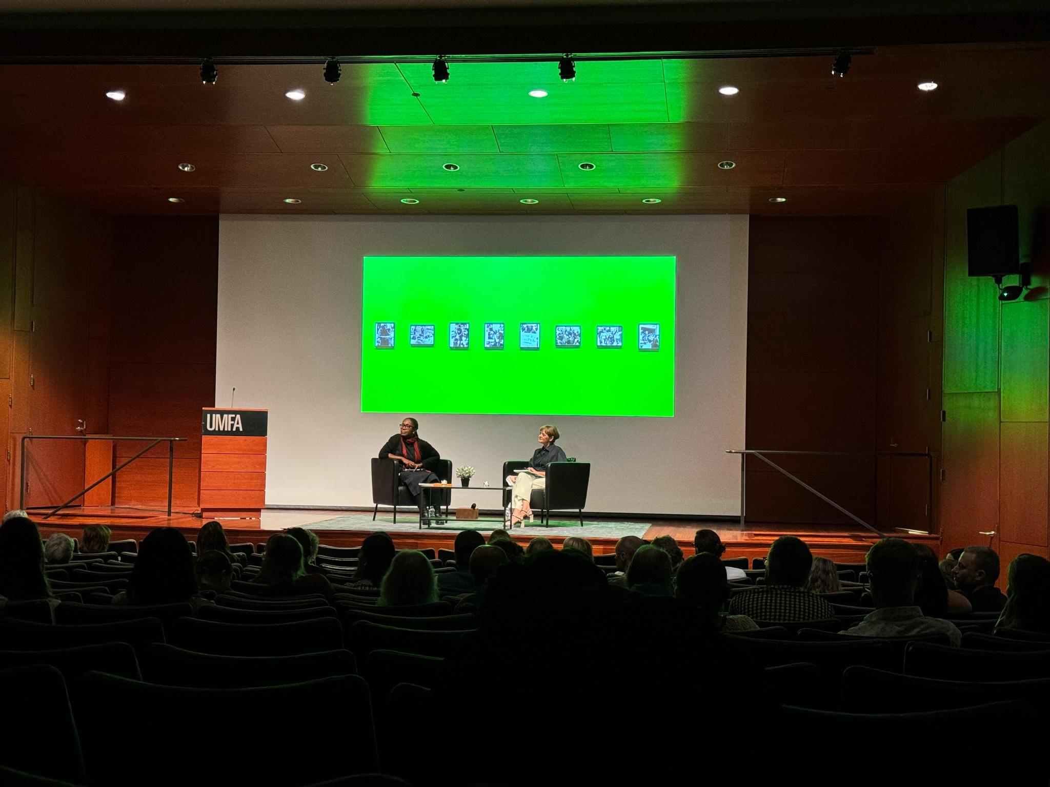 two people sitting on a stage with a projected green image behind them and with a crowd in the foreground