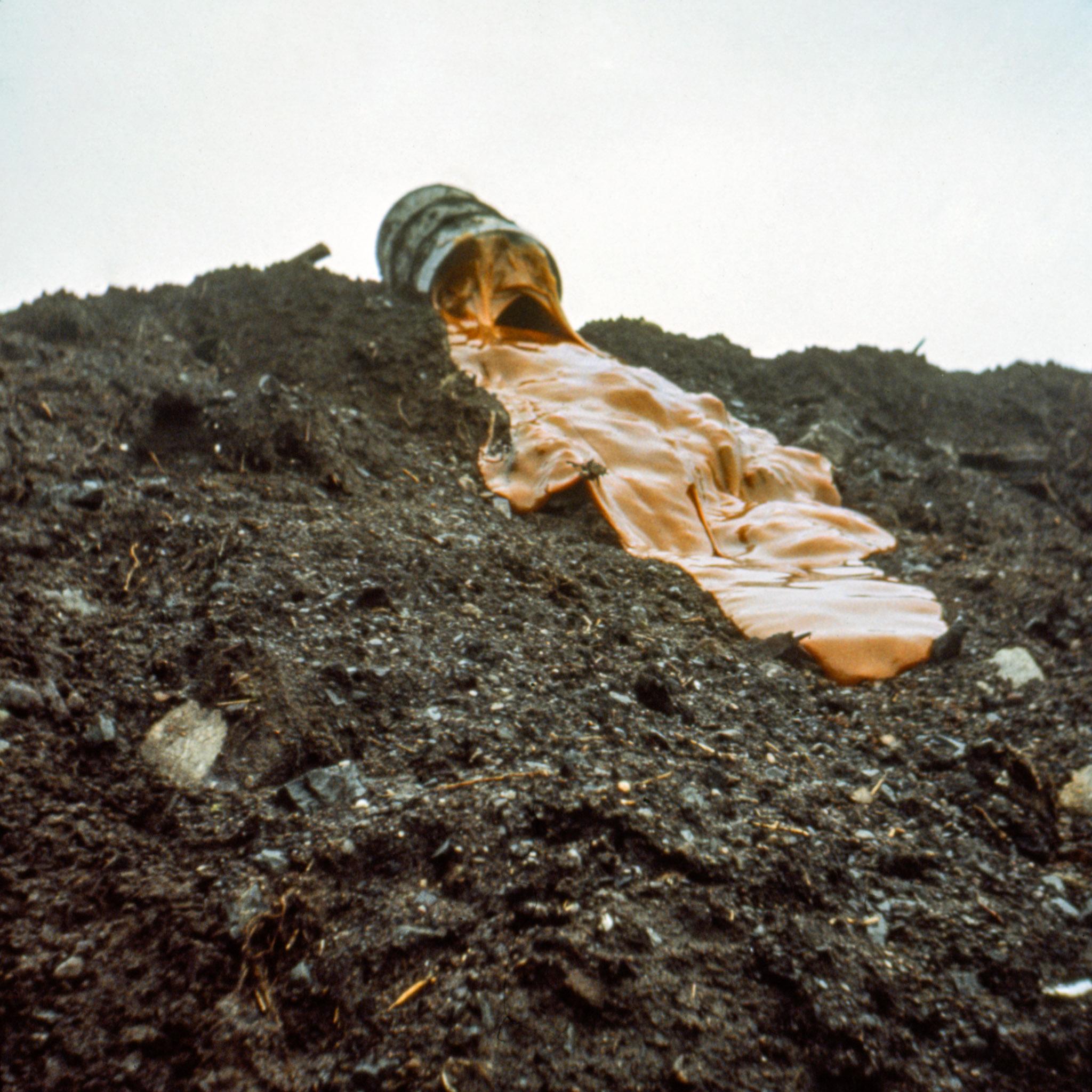 Viscous orange glue slowly pouring down a muddy hillside out of a large barrel