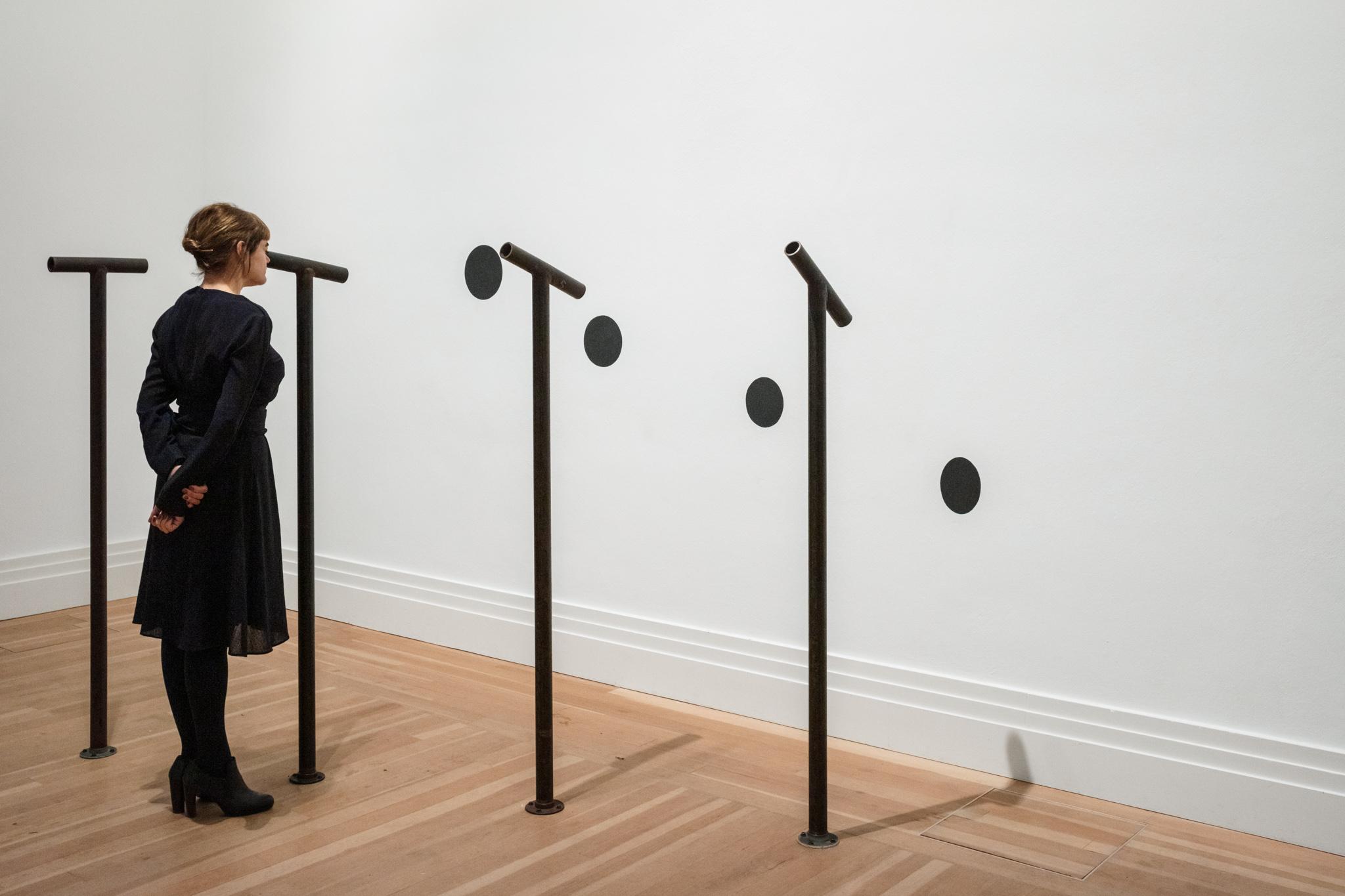 a person looking through one of four steel pipes attached to the floor with viewing pipes attached to the top, each looking at a painted black circle on a wall