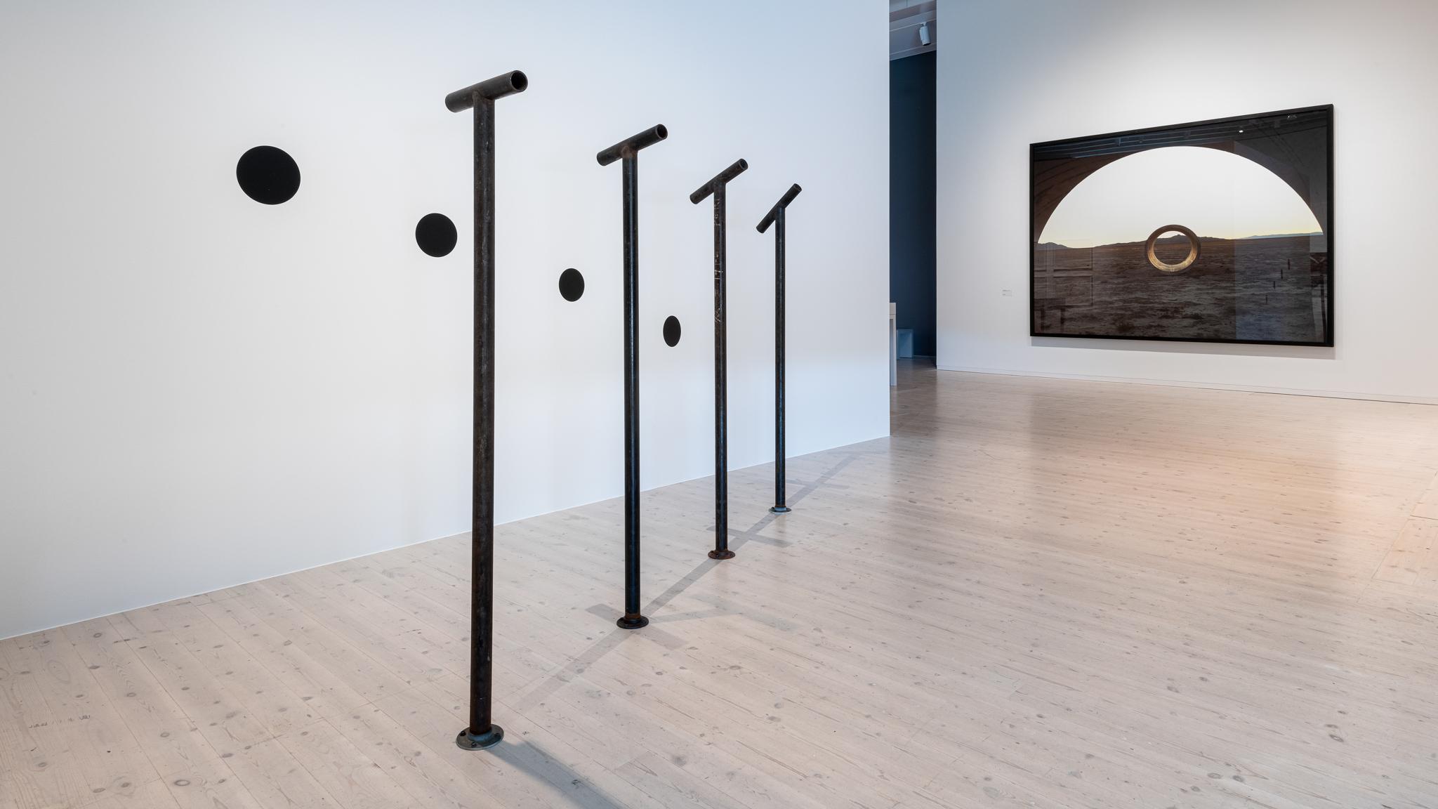 four steel pipes attached to the floor with viewing pipes attached to the top, each looking at a painted black circle on a wall