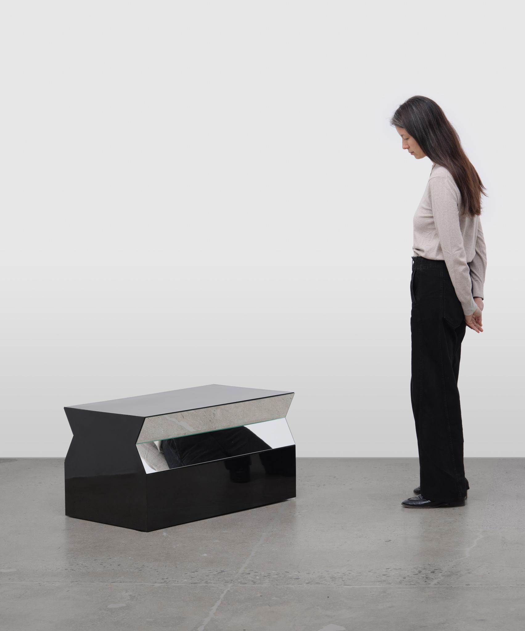 a figure looking at a geometric sculpture made of formica and mirrors sitting on a concrete floor