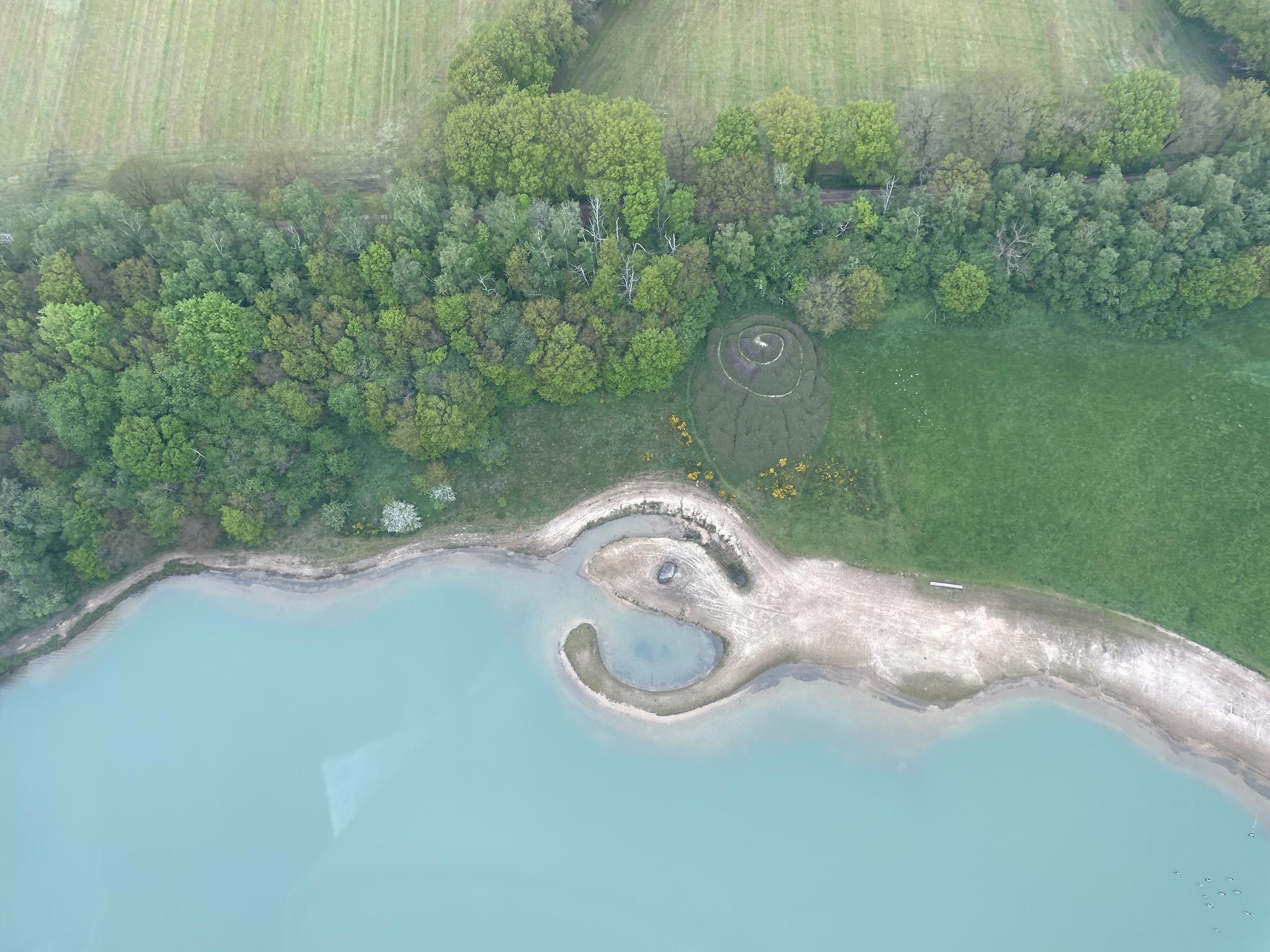 Broken Circle / Spiral Hill, a circular earthwork extending into blue water with a green hill with spiral trail on the shore, pictured in 2021 from above