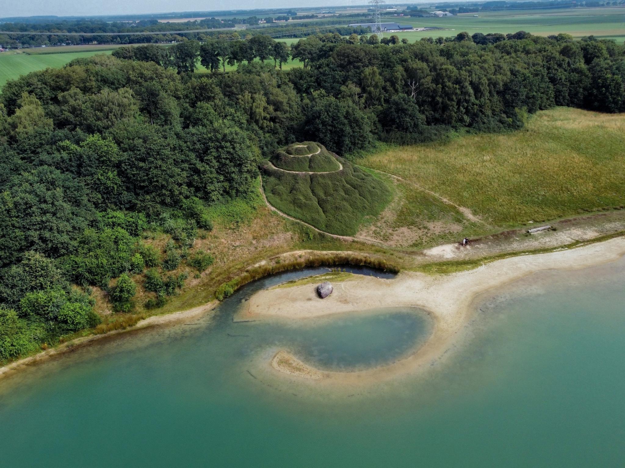 Broken Circle / Spiral Hill, a circular earthwork extending into blue water with a green hill with spiral trail on the shore, pictured in 2025 from above